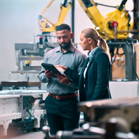 Person writing on paper, another holding phone, laptop and coffee cup on table.
