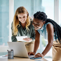 Two women leaning over a laptop, reviewing documents and the screen.