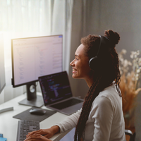 Woman wearing headphones, smiling while working on a laptop and monitor in front of a sunlit window.