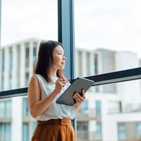 Young woman holding a tablet, gazing out of a window. She appears to be deep in thought or reflecting