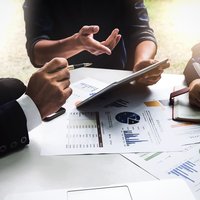People in a business meeting discussing financial charts, taking notes, and using a tablet at a sunlit table.