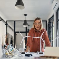 A young woman in professional attire standing around a desk, engaged in discussion. On the desk are model wind turbines.